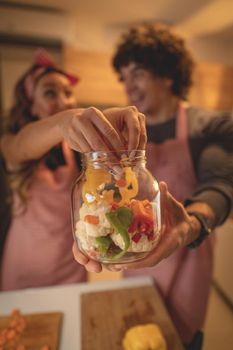 Cute couple is putting vegetables in a jar and smiling while making pickle in kitchen at home