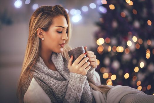 Cute young woman holding cup of coffee and sitting in the bed with Christmas lights. 