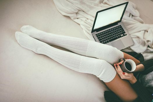 Close-up of the young woman's legs sitting on the bed with laptop and cup of coffee in hands.