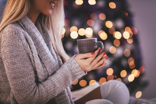 Cute young smiling woman sitting in the bed and holding cup of coffee or tea, surrounded with Christmas lights. 
