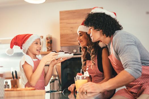 Happy parents and their daughter with santa's cap are preparing meal together in the kitchen. Little girl is holding plate with muffin offering to her mother and father.