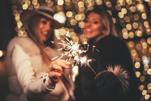 Young beautiful cheerful female friends with sparklers having fun and enjoying in the city street at Christmas eve. Selective focus. Focus on foreground, on sparklers.