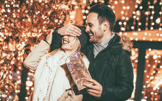 Man keeps his girlfriend eyes covered while she giving a gift for Christmas holiday.