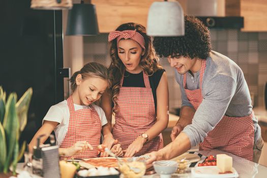 Happy parents and their daughter are preparing meal together in the kitchen while little girl is putting ketchup on the pizza dough.