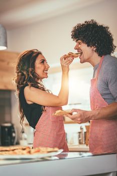 Happy couple enjoys and having fun in their home kitchen. Young woman feeds her loving man with piece of pizza.