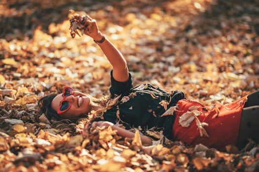 Beautiful young woman is enjoying in autumn  sunny forest colors. She is lying down on golden yellow leaves and having fun. 