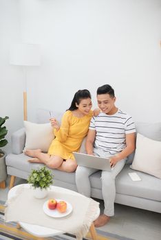 Pretty Asian woman and handsome man relaxing on couch with laptop and surfing Internet while making purchases online