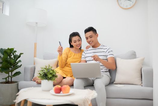 Pretty Asian woman and handsome man relaxing on couch with laptop and surfing Internet while making purchases online