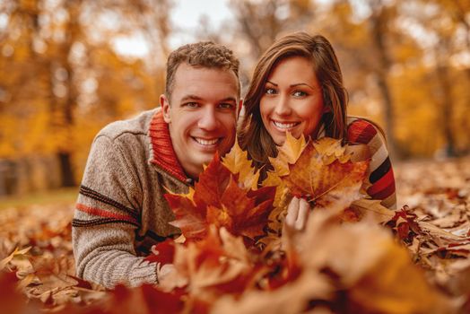 Beautiful young couple in sunny forest in autumn colors. They are lying on the ground holding fallen leaves and enjoying. 