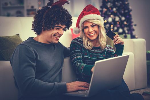 Cheerful young couple relaxing at home and online buying Christmas presents on laptop.