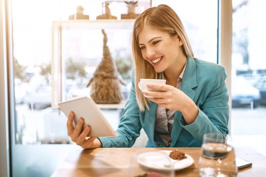 Beautiful young smiling woman drinking coffee in a cafe. She is surfing the internet on a digital tablet.