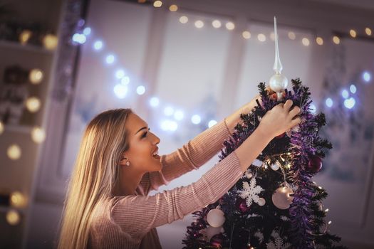 Beautiful smiling young woman decorating christmas tree with Christmas ornament at home.