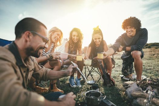 Happy young friends enjoy a sunny day at the mountain. They're laughing and grilling -roasting sausages on sticks over a campfire near tent.