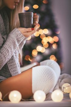 Close-up of a cute young woman sitting in the bed and holding cup of coffee or tea, surrounded with Christmas lights. 