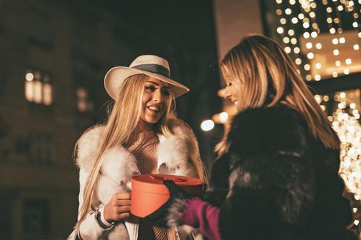 Young beautiful two cheerful sisters are celebrating Christmas on the city street, giving presents to each other. 