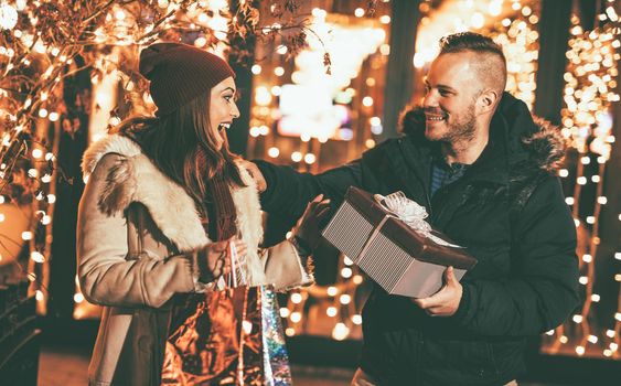 Young beautiful cheerful couple celebrating Christmas in the city street and giving many gift to each other. 