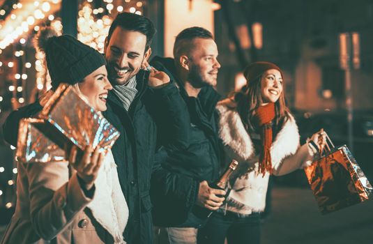 Four smiling young friends standing outside and holding many shopping paper bags and boxes in city holiday night. Selective focus. Focus on foreground.