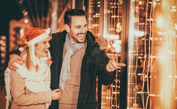 Couple having fun outdoors at Christmas time pointing at something behind shop window.