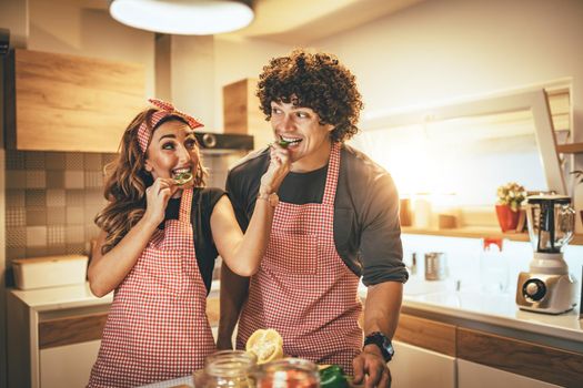 Happy young couple enjoys and having fun in eating slices of capsicum and making healthy meal together at their home kitchen.