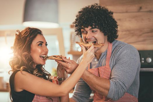 Happy couple enjoys and having fun in their home kitchen. Young man and woman feed each other with pieces of pizza.