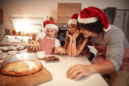 Happy parents and their daughter with santa's cap are preparing meal together in the kitchen. Little girl is offering lunch to her mother and father.