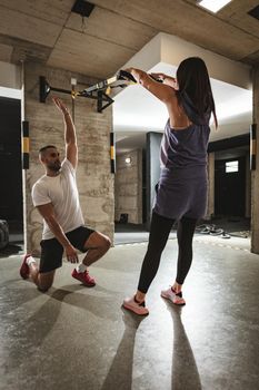 Rear view of young handsome woman having workout with a suspension straps at the gym supported of her personal male trainer. 