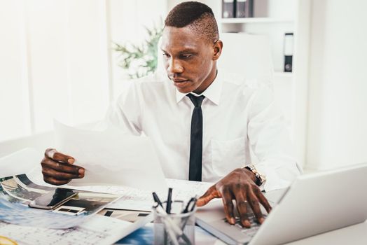 Successful  African businessman looking on paper document and working on laptop in modern office.