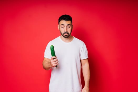 Young man in a white shirt isolated over background. Guy look at long cucumber in hand.