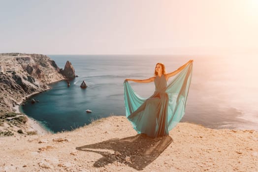 Side view a Young beautiful sensual woman in a red long dress posing on a rock high above the sea during sunrise. Girl on the nature on blue sky background. Fashion photo.