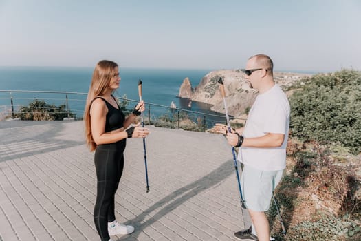 Happy Middle aged couple or friends practicing nordic walking in park near sea. Mature couple with trekking poles walking, practicing Nordic walking outdoors. Aging youthfully and sport concept.