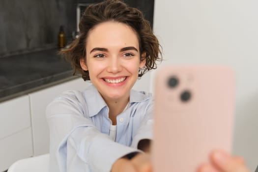 Portrait of beautiful young woman, taking selfie on mobile phone at home, poses for photo with smartphone.