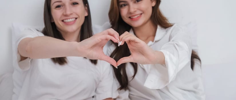 Lesbian couple together concept. Portrait of a smiling young lesbian couple other while sitting together and hand made heart symbol in the morning at bedroom.