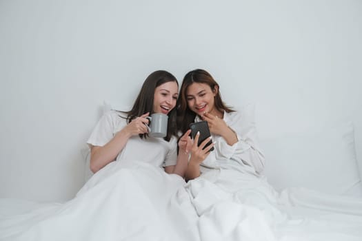 Two Asian cheerful happy female girlfriends having a coffee sitting on the bed while looking at the smartphone. Concept lifestyle lesbian couple.