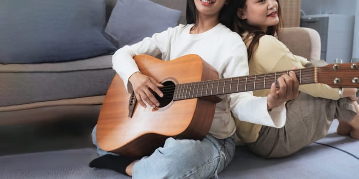 Homosexual LGBT young women playing guitar back against each other and singing in living room with happiness moment.