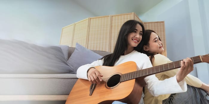 Homosexual LGBT young women playing guitar back against each other and singing in living room with happiness moment.