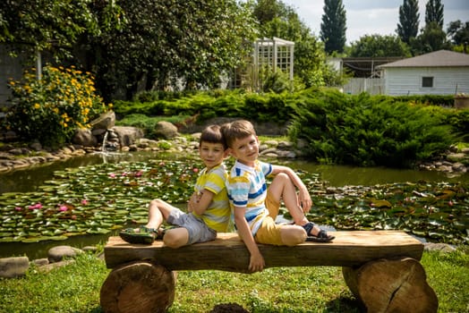 Two brother boys sitting on a bench in a park near peaceful lake with water lily Nymphaeum. Kids relaxing on nature on hot summer day.