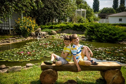 Two brother boys sitting on a bench in a park near peaceful lake with water lily Nymphaeum. Kids relaxing on nature on hot summer day.