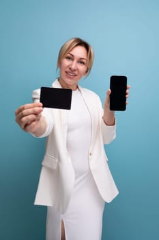 confident young european woman in a white jacket and dress advertises a bank card and a smartphone.