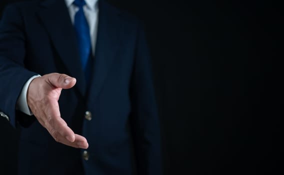 Businessman standing and doing handshake gesture on dark background. Business concept. Copy space.