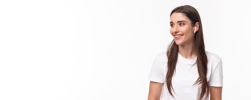 Close-up portrait of lovely, pretty young woman in t-shirt, listening music in wireless headphones, look away with pleased smile, enjoying favorite podcast or songs playlist, white background.