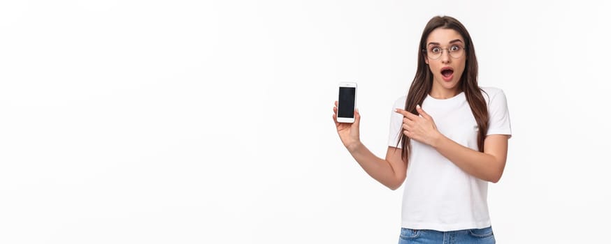 Communication, technology and lifestyle concept. Portrait of impressed and excited young brunette girl in glasses, pointing at mobile phone display with gasp and amazed face, white background.