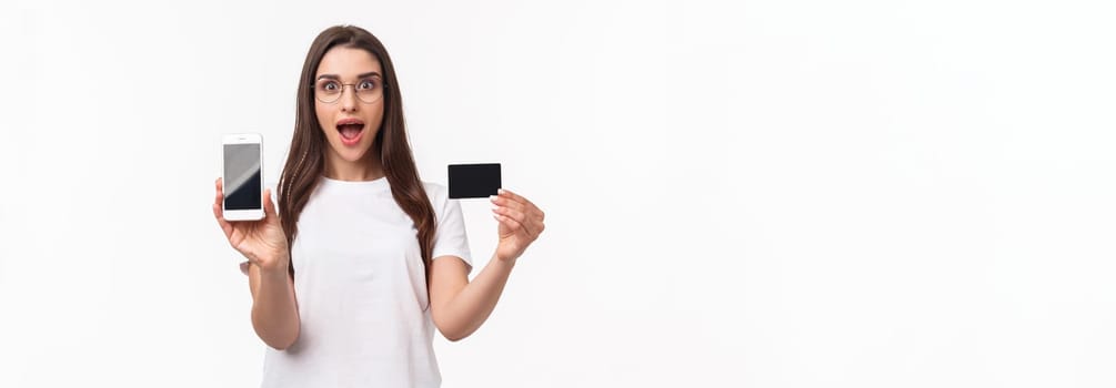 Portrait of excited and happy young woman introduce new application, favorite online store to buy clothes, show mobile phone and credit card, smiling amused, white background.