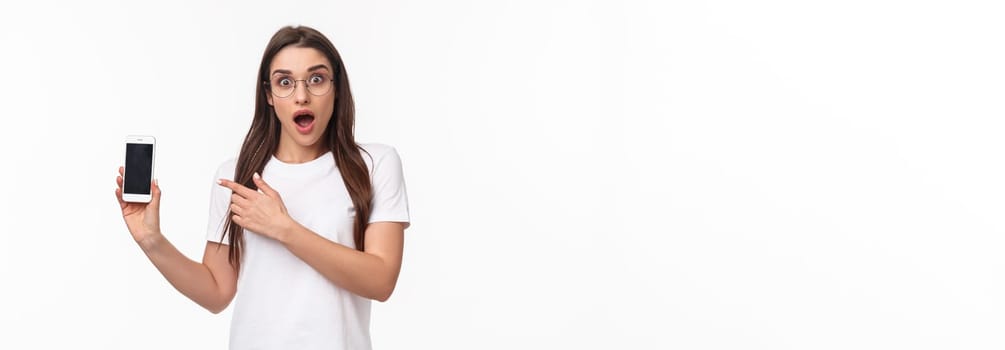 Communication, technology and lifestyle concept. Portrait of impressed and excited young brunette girl in glasses, pointing at mobile phone display with gasp and amazed face, white background.