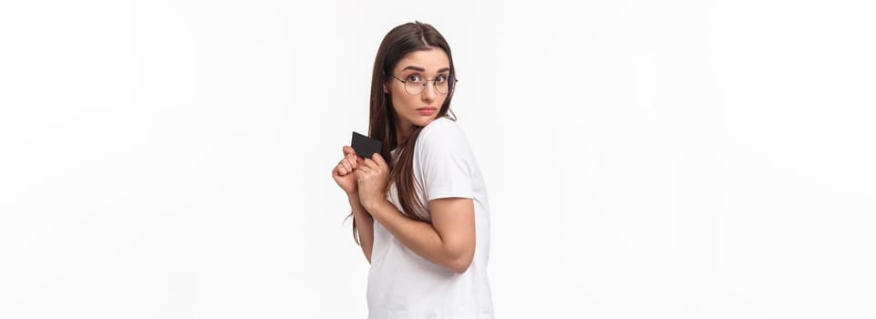Portrait of greedy and funny young woman unwilling to give her credit card, hiding it and looking with disbelief and reluctance, have money but dont like share, standing white background.