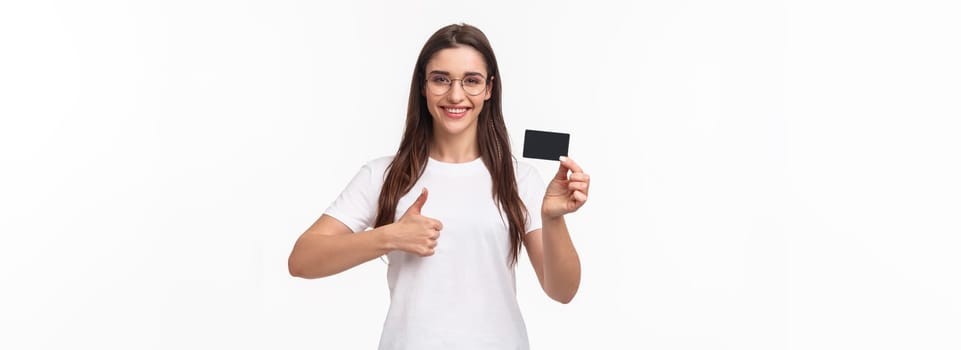 Portrait of pleased, attractive young brunette woman in t-shirt and glasses, recommend bank, showing credit card, smiling satisfied show thumbs-up, approve like and advice make online purchases.