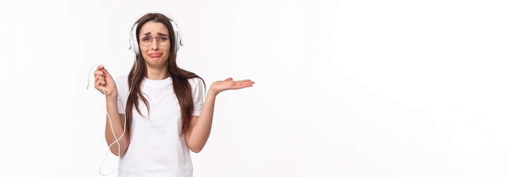 Technology, lifestyle and music concept. Portrait of awkward and frustrated young gloomy girl in headphones, holding wire and shrugging upset, earphones broke, want buy wireless earbuds.