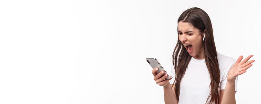 Close-up portrait of pissed-off aggressive and angry young brunette woman yelling at person while having phone call in wireless earphones, screaming at smartphone outraged.