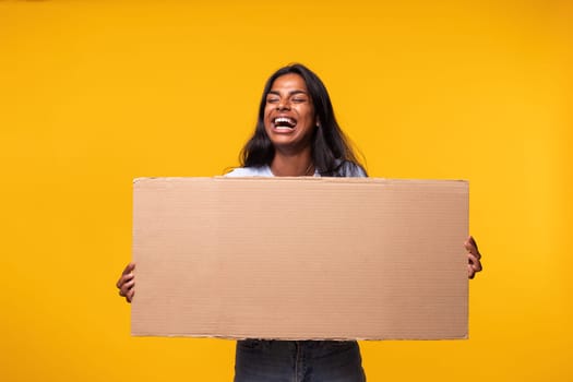 Young Indian woman laughing holding a cardboard banner isolated in yellow background. Studio shot. Activist concept.