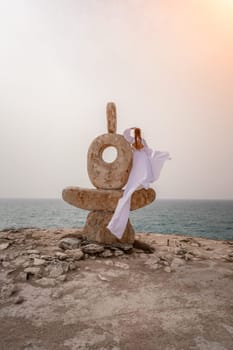 Woman sea stone. A woman stands on a stone sculpture made of large stones. She is dressed in a white long dress, against the backdrop of the sea and sky. The dress develops in the wind