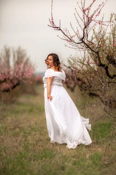 Woman peach blossom. Happy woman in white dress walking in the garden of blossoming peach trees in spring.